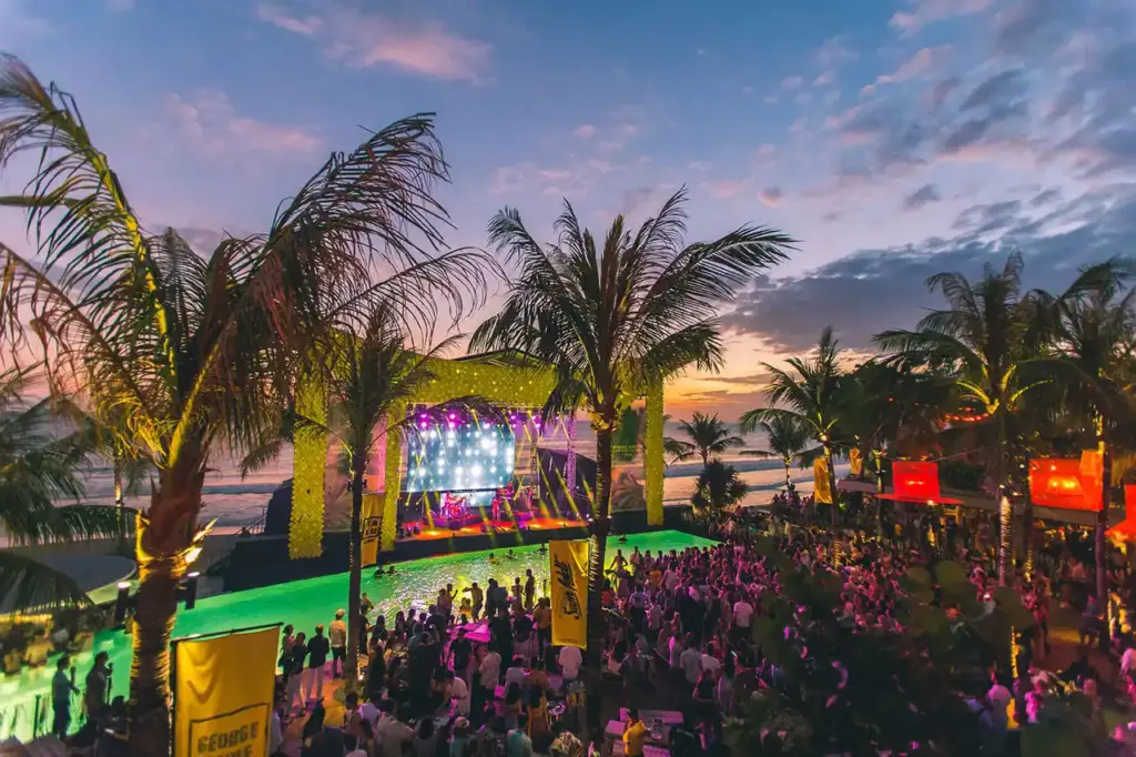 Crowds gathering at a beachfront stage with stage lights and palm trees at sunset, capturing the lively nightlife scene beyond the bars in Ubud.