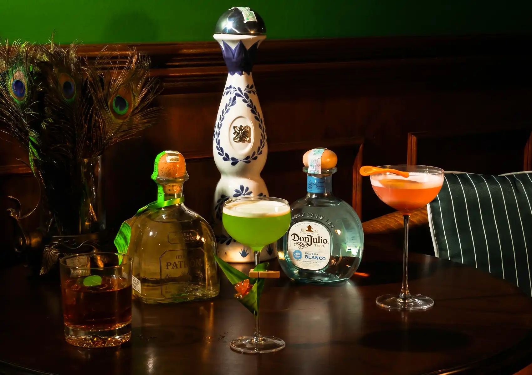 A dramatic bar display featuring Patrón and Don Julio Blanco tequila bottles flanking a tall ornate blue-and-white ceramic mezcal decanter, surrounded by three craft cocktails — an amber drink in a rocks glass, a frothy green cocktail in a coupe, and a vibrant orange drink in a tall martini glass garnished with a citrus twist — set against a dark wood backdrop with peacock feathers on the left