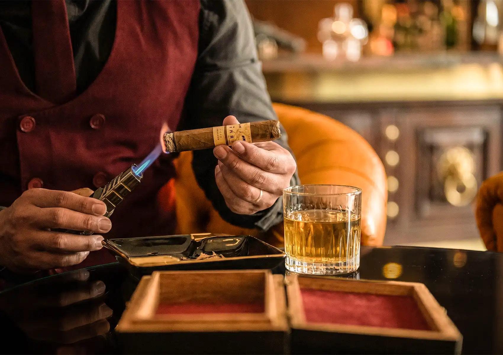 A uniformed cigar sommelier in a burgundy vest lighting a premium hand-rolled cigar with a blue-flame torch lighter over a black ashtray, with a crystal rocks glass of whisky and an open red-velvet cigar box on the table and a warmly lit bar in the background