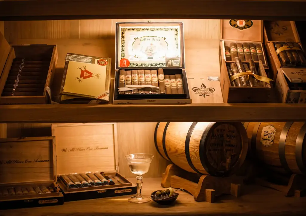 Warmly lit humidor shelves displaying an extensive collection of premium cigars in open wooden boxes including a featured Bellas Artes box rated #1, alongside a small oak aging barrel, a martini glass with olives, and cigar boxes inscribed with "We All Have Our Reasons"