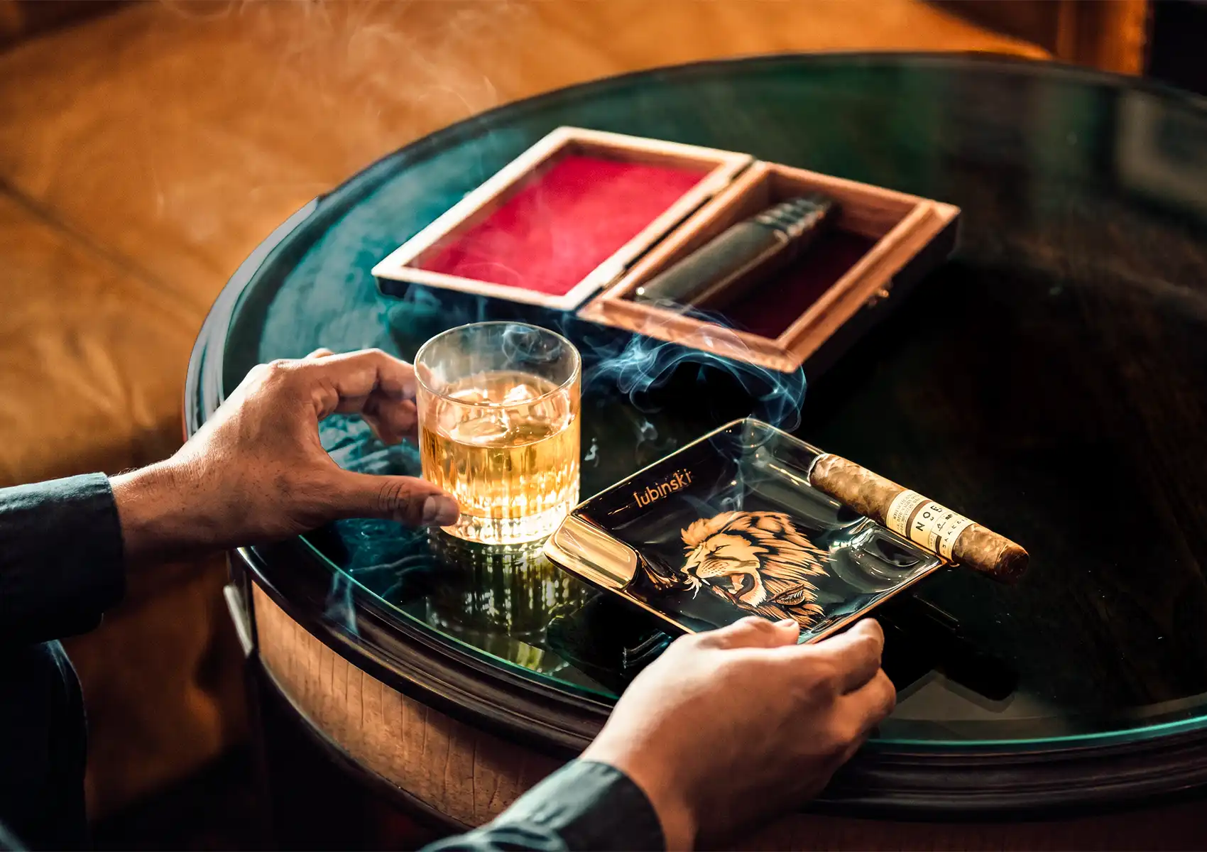 A person holding a rocks glass of whisky over a glass table while another hand holds a Lubinski lion-emblazoned ashtray with a lit cigar resting on it, with wisps of blue smoke rising and a red-velvet-lined wooden cigar box open in the background