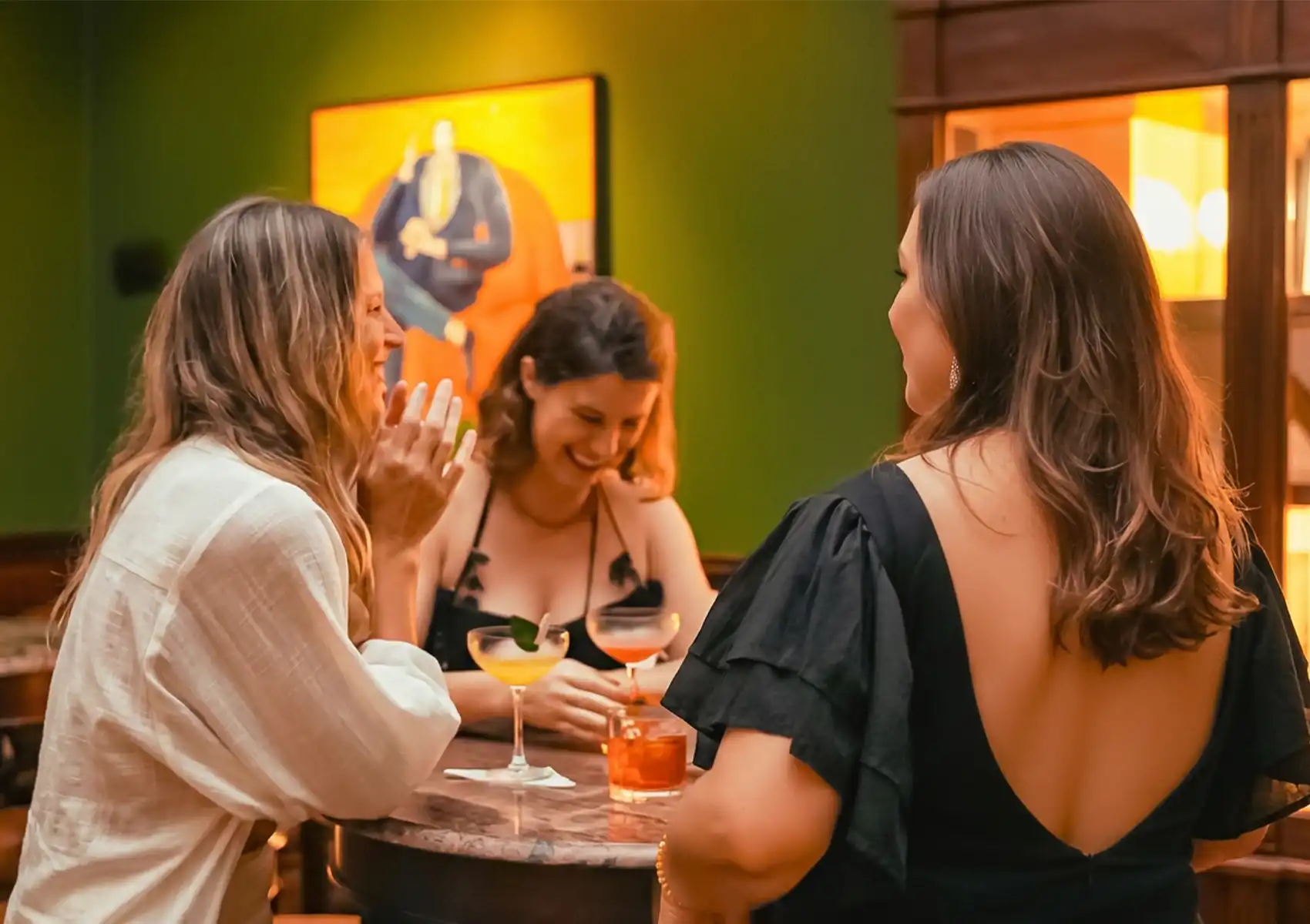 Friends enjoying cocktails in a stylish Ubud lounge, gathered around a small table with warm lighting and vibrant artwork on the wall.