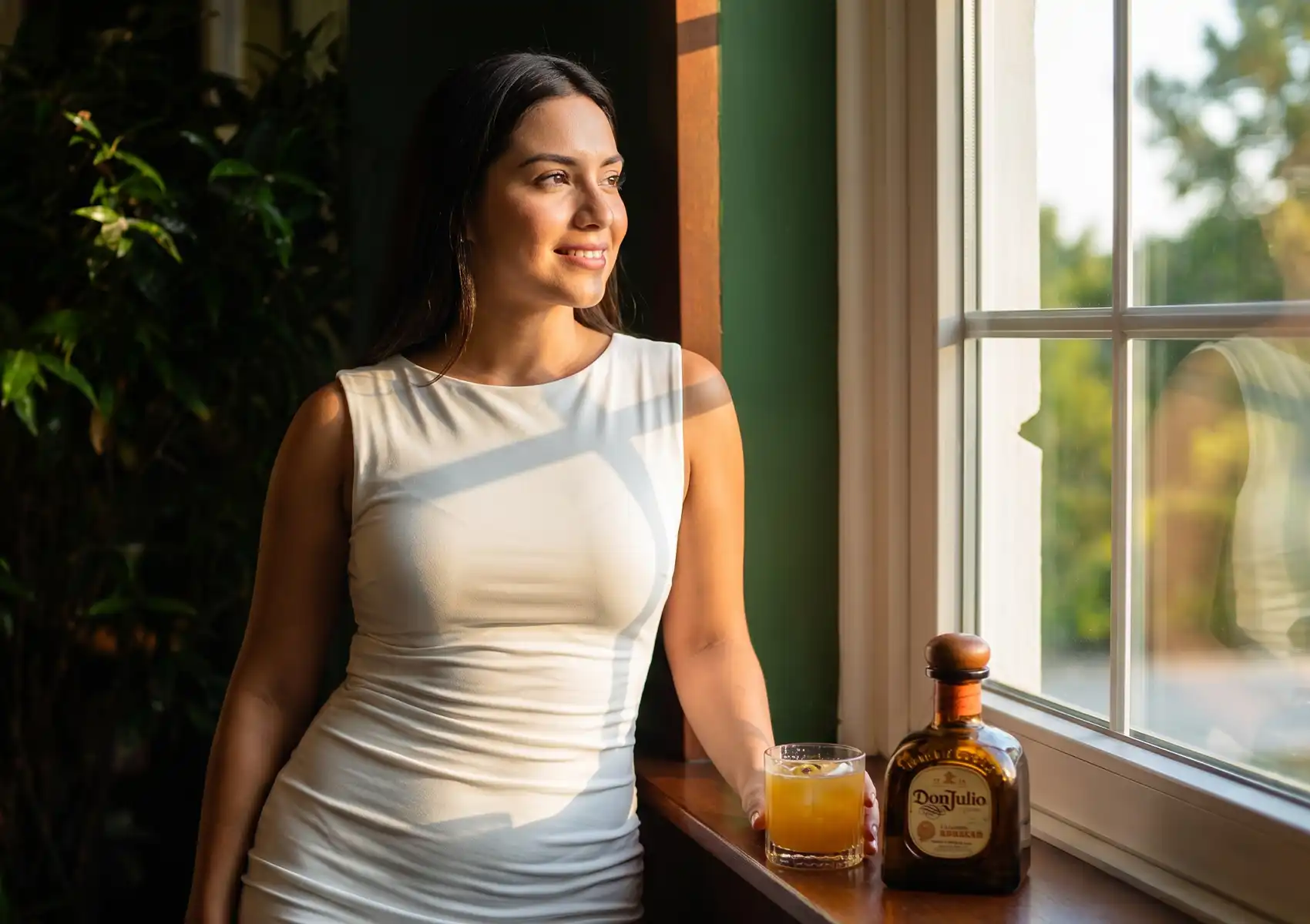 A smiling woman in a white sleeveless dress bathed in warm golden hour sunlight, leaning against a windowsill beside a rocks glass of orange cocktail and a bottle of Don Julio tequila, with lush green tropical foliage visible through the window