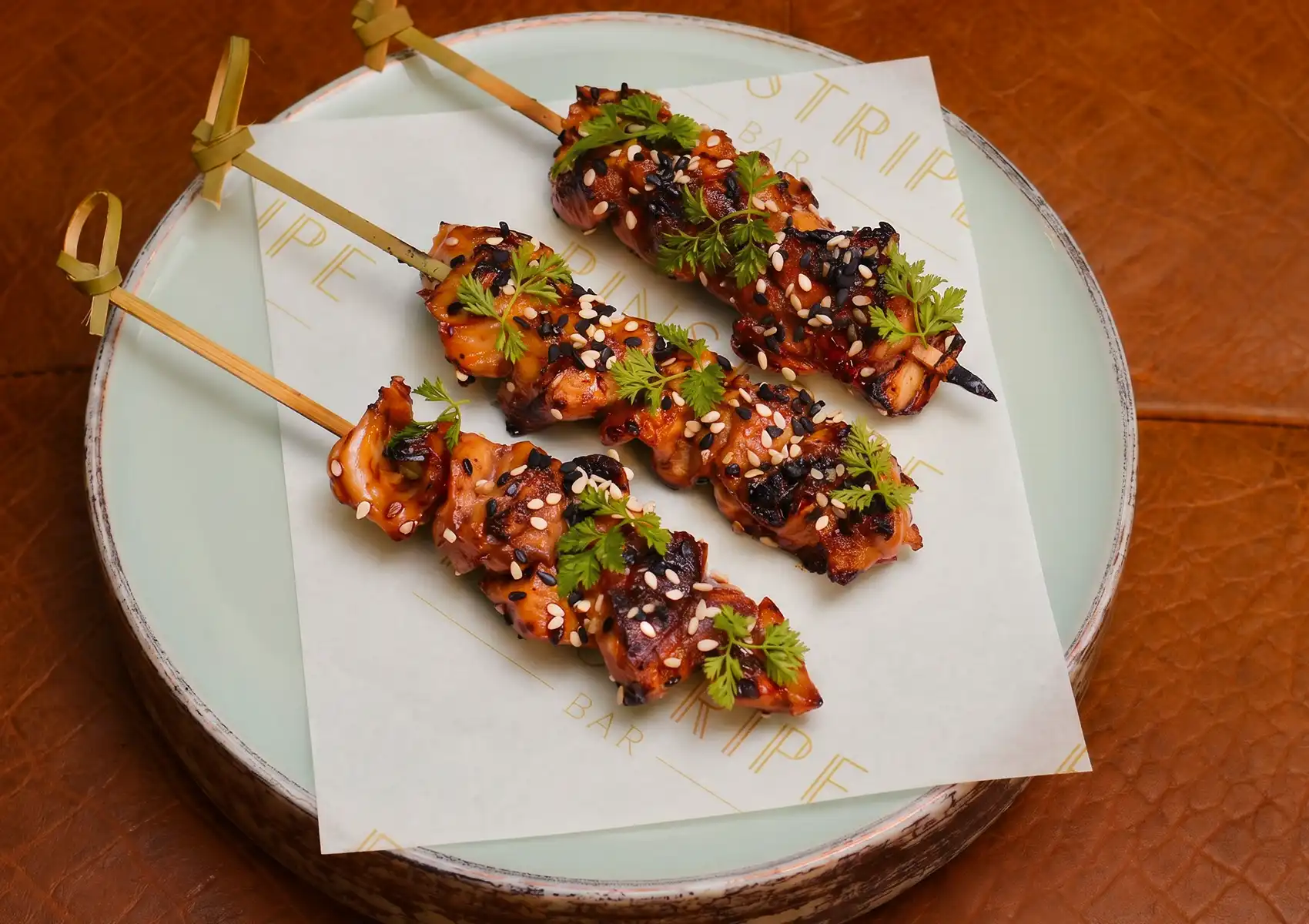 A close-up, overhead view of three artisan yakitori skewers coated in a rich glaze, sprinkled with black and white sesame seeds, and topped with fresh green herbs, making them an ideal gourmet snack to enjoy at a lively cocktail bar ubud.