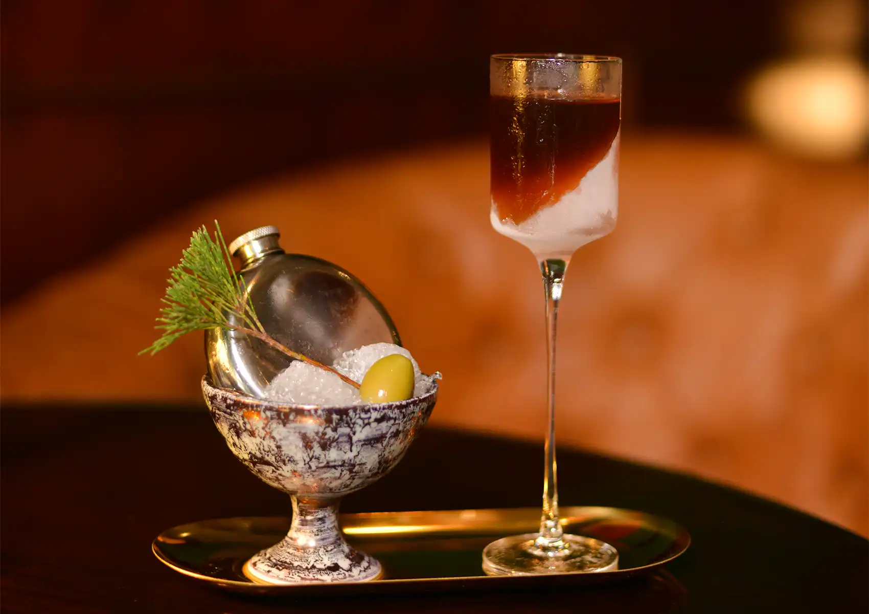 An ornate and creative cocktail presentation on a brass tray, featuring a tall glass with a layered dark and light drink next to a decorative bowl holding crushed ice, a small silver flask, a pine sprig, and a green olive, highlighting the unique mixology at a sophisticated cocktail bar ubud.