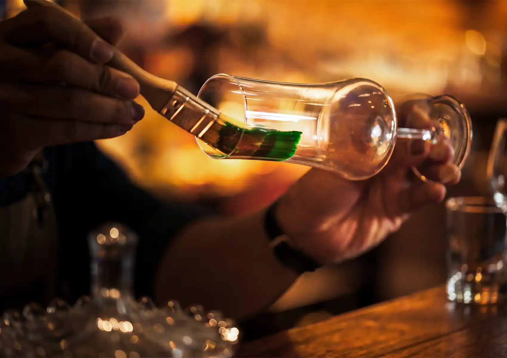 A mixologist's hands meticulously using a small brush to paint a bright green, edible streak inside a clear stemmed glass, showcasing the highly artistic drink preparation at a sophisticated Cocktail Bar Ubud.