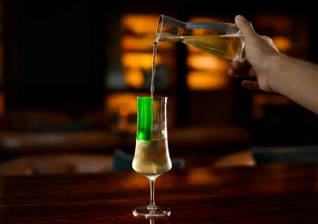 A bartender smoothly pouring a crafted golden drink from a glass carafe into a custom-painted tall glass resting on a warm, polished wooden counter at a fine Cocktail Bar Ubud.