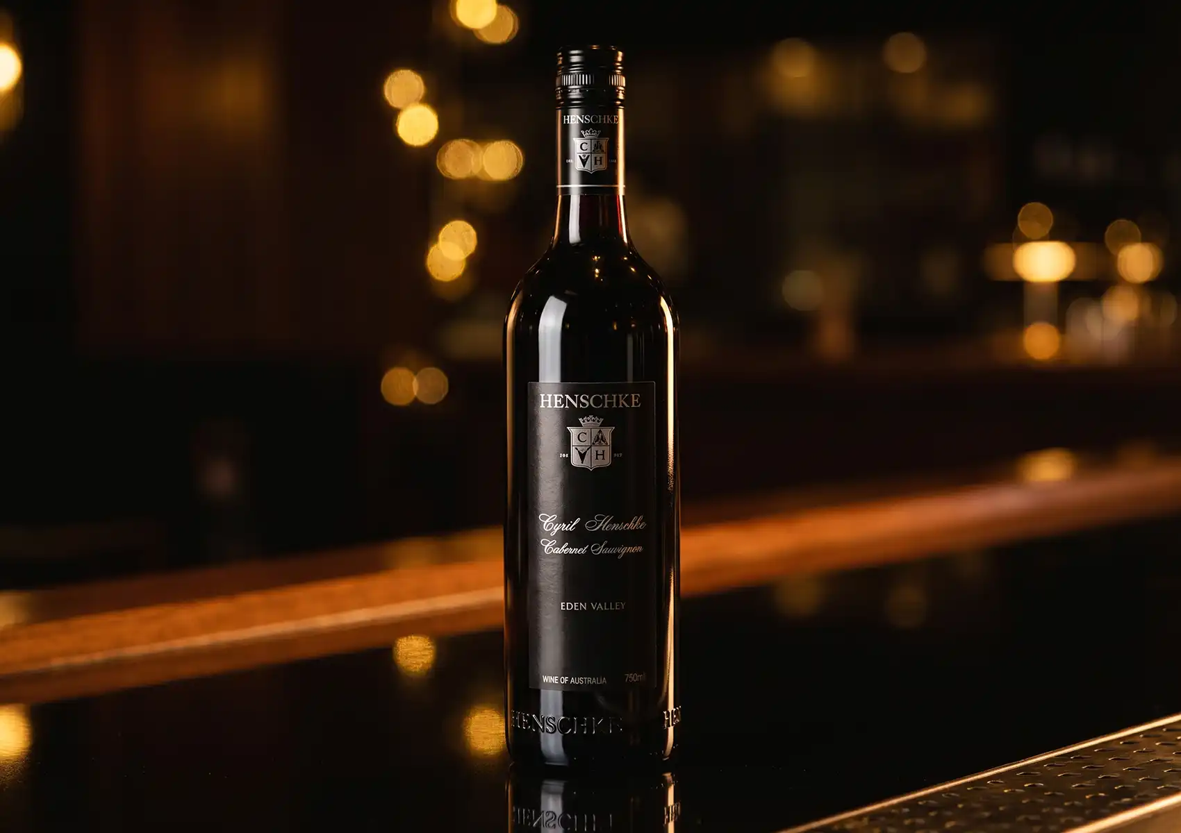 A sleek bottle of Henschke Cyril Henschke Cabernet Sauvignon resting on a polished, reflective counter with warm, blurred bokeh lights in the background, showcasing the fine beverages available at a premier cocktail bar ubud.