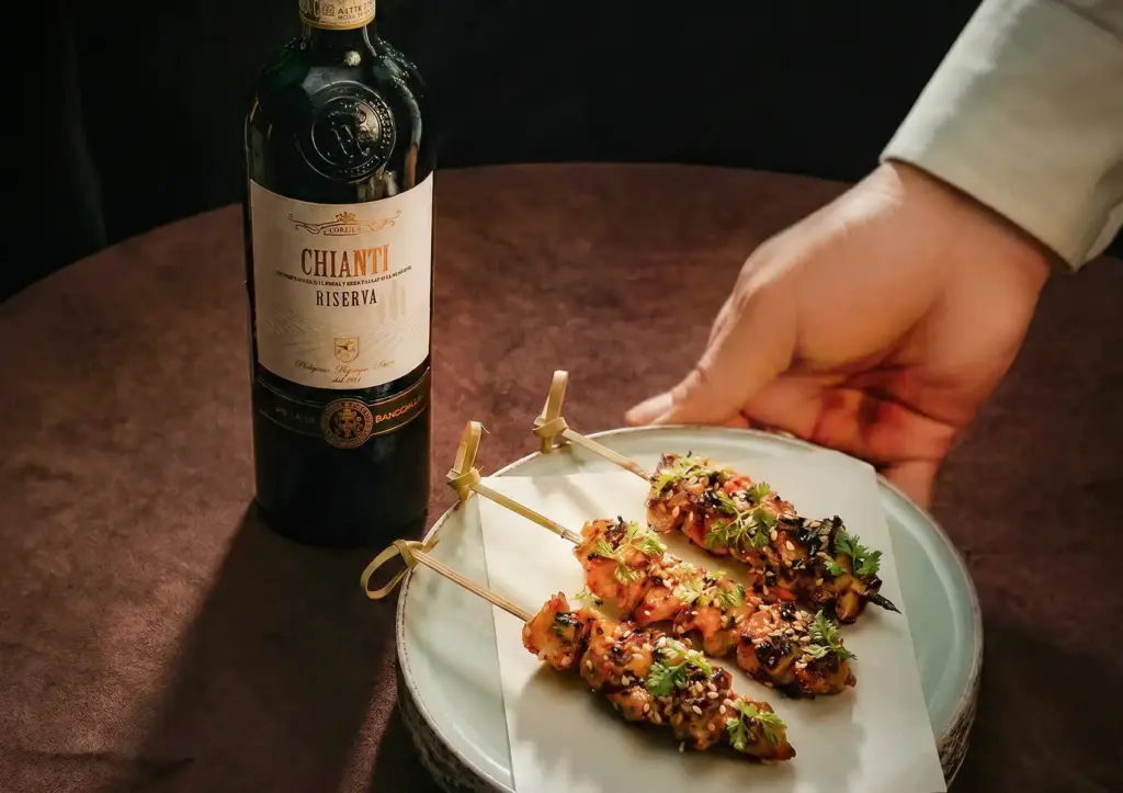 A server's hand placing a plate of three beautifully glazed yakitori skewers garnished with sesame seeds and herbs next to a bottle of red wine, showcasing the delicious culinary pairings available at a premium cocktail bar ubud.