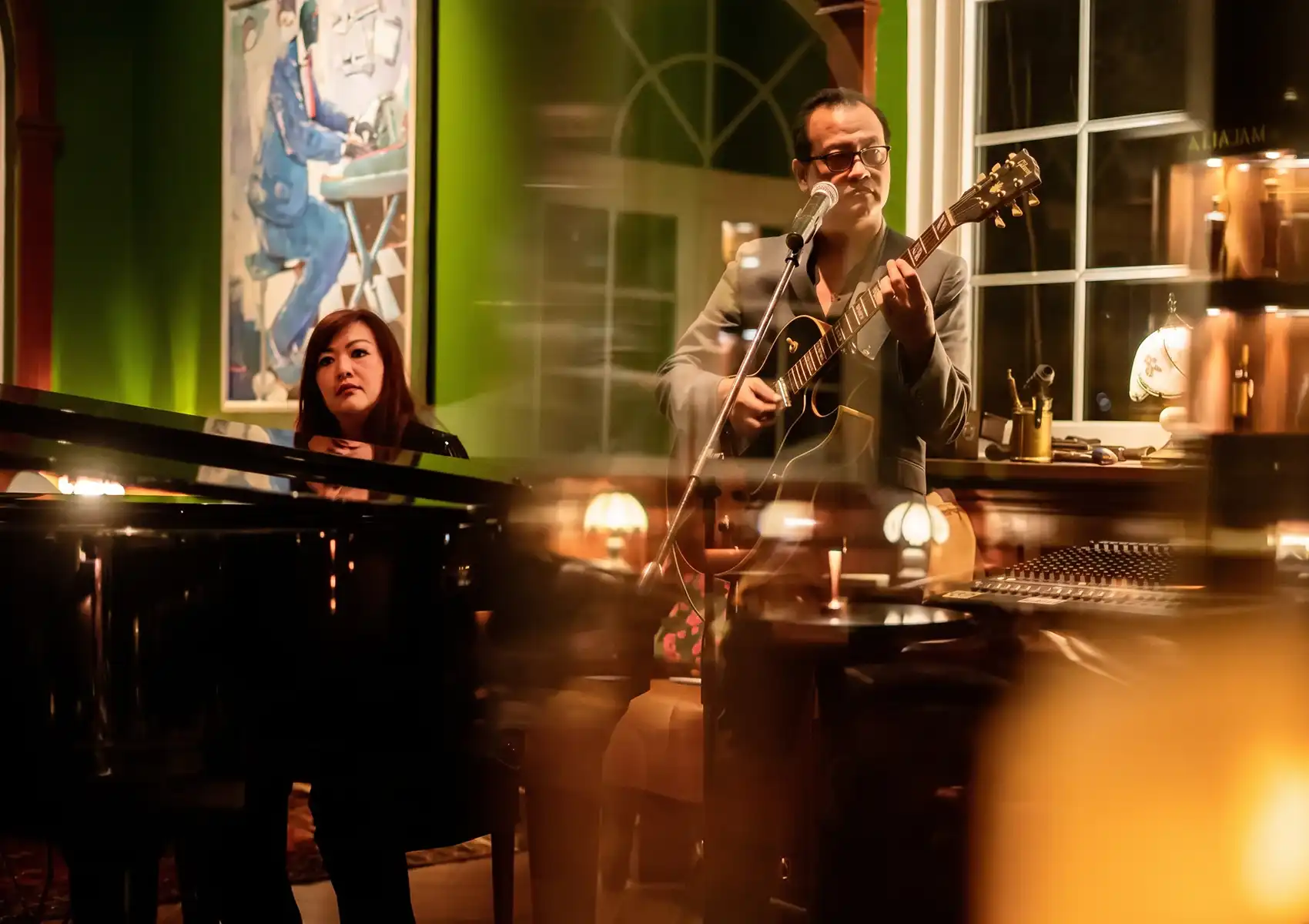 A man playing an acoustic guitar and singing at a microphone alongside a woman playing a black grand piano in a warmly lit, elegantly decorated green room, providing live entertainment for guests at a stylish cocktail bar ubud.