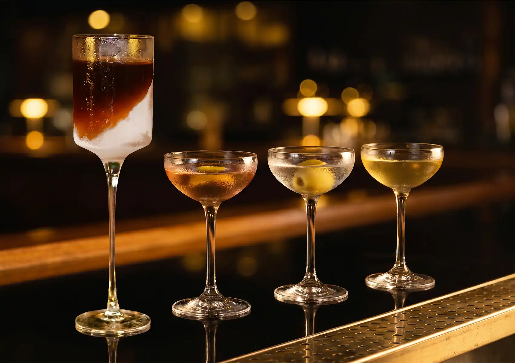 A beautiful lineup of four elegant drinks resting on a sleek, reflective counter, featuring a tall, striking layered beverage and three classic coupe glasses with various garnishes, showcasing the vibrant drink selection at a premier cocktail bar ubud.
