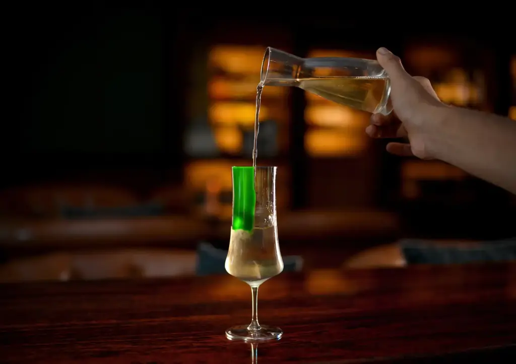 A bartender pouring a light-colored mixed drink from a glass carafe into an elegant stemmed glass featuring a bright green garnish at an upscale cocktail bar Ubud.