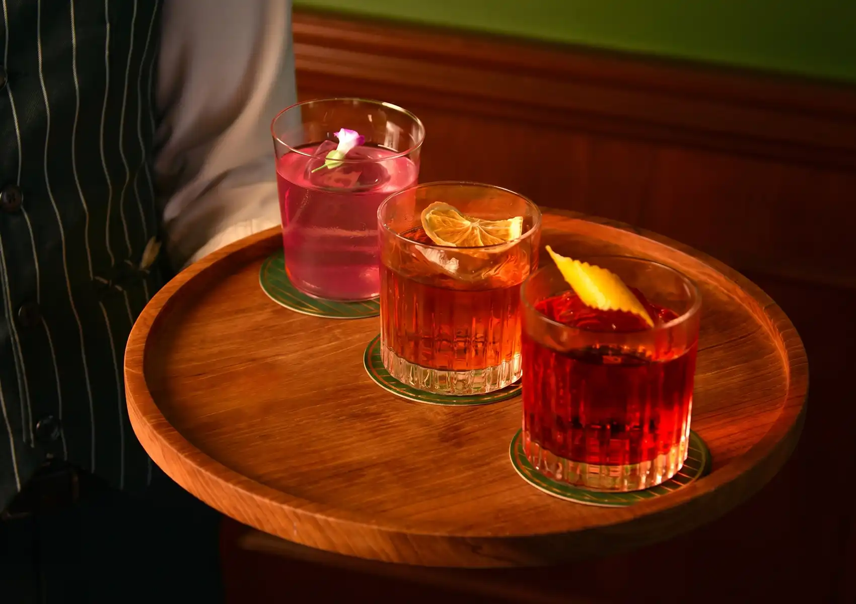 A server holding a round wooden tray presenting a flight of three vibrant, beautifully garnished drinks served in a stylish, dimly lit cocktail bar Ubud.