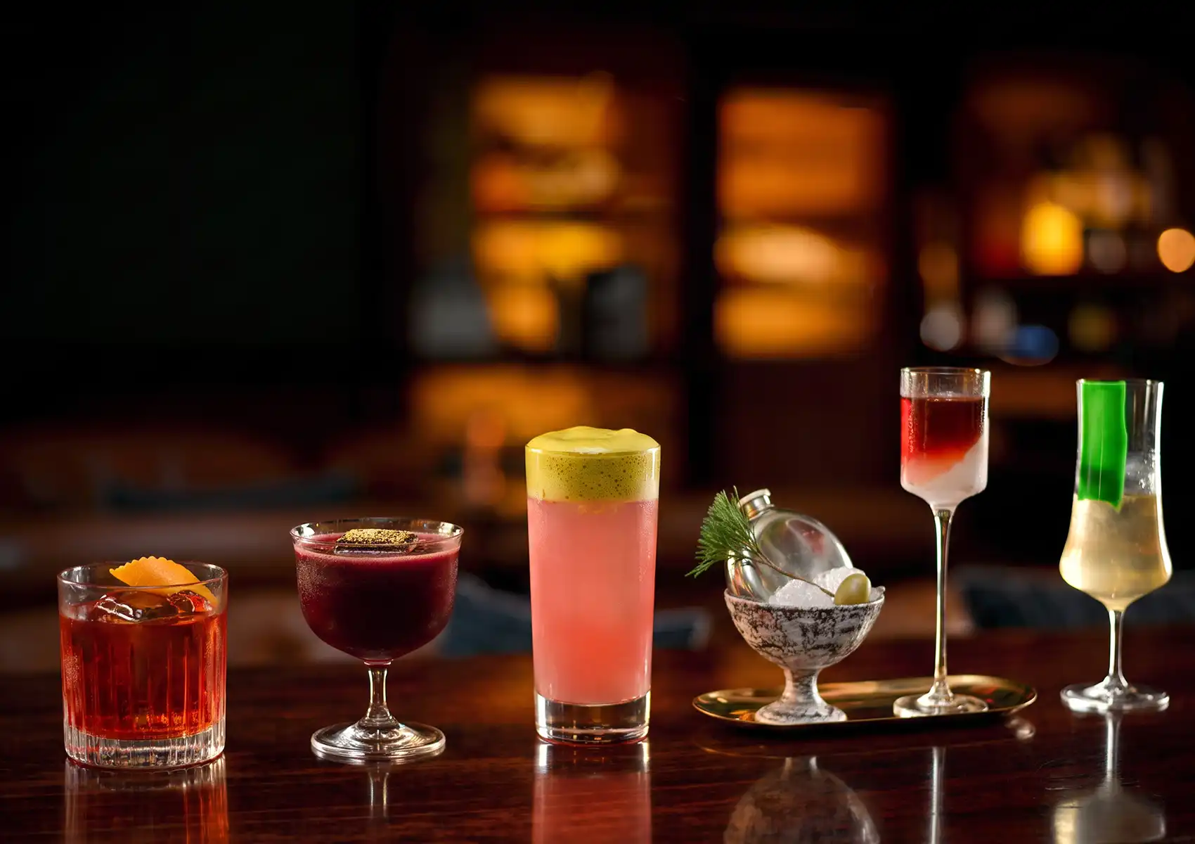 A vibrant lineup of six uniquely crafted, colorful drinks presented in diverse and elegant glassware on a dark wooden counter at a cocktail bar Ubud.