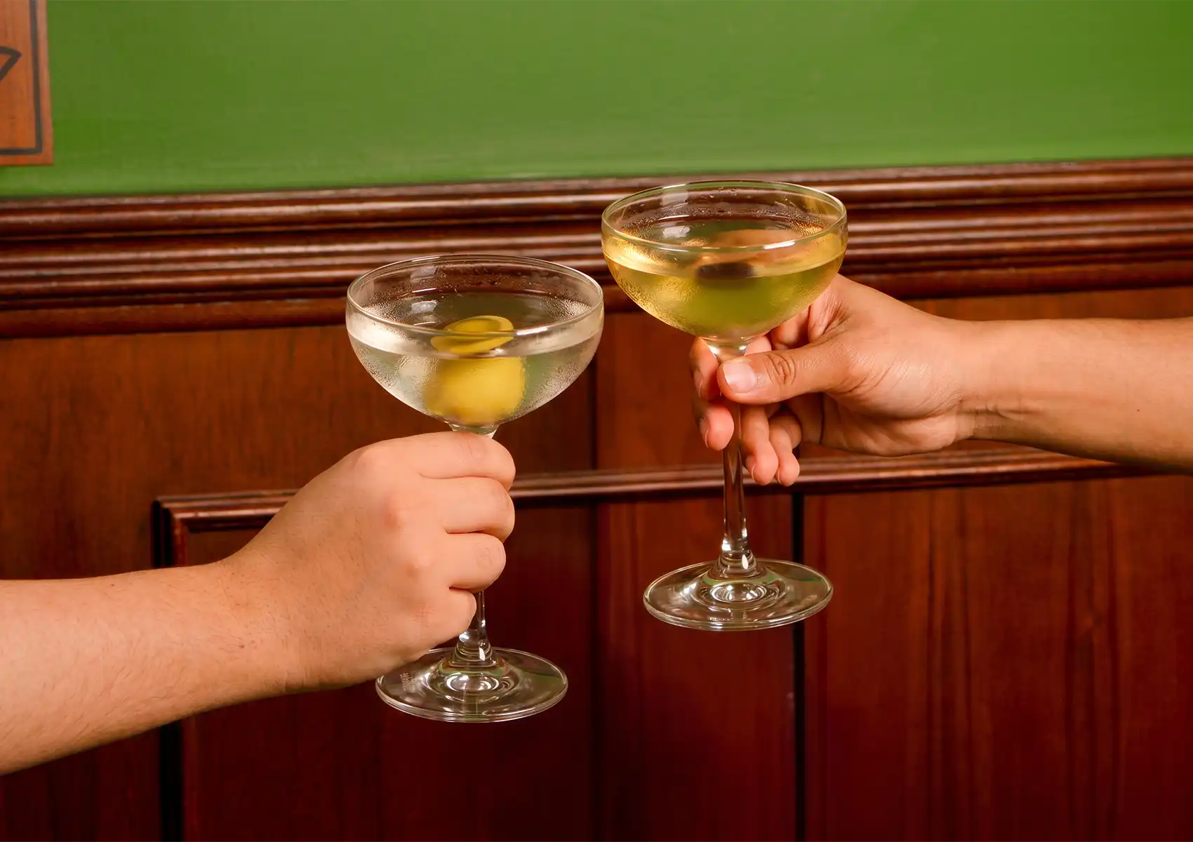 A close-up of two people clinking their glasses together in a toast, enjoying some of the best martini cocktails featuring a classic olive garnish, set against a warm wooden and green background.