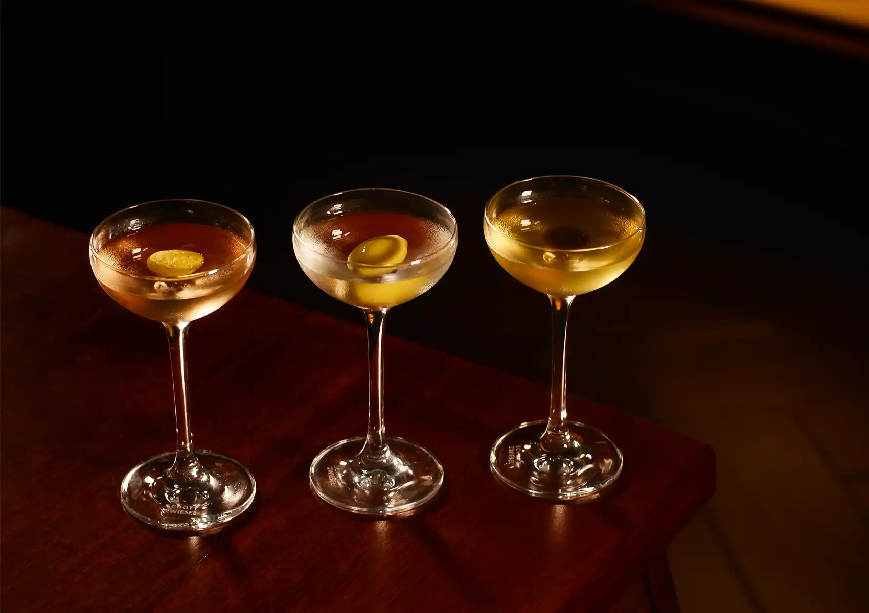 Three elegant coupe glasses resting on a polished dark wooden table, showcasing a beautifully crafted flight of the best martini cocktails in a moody, atmospheric bar setting.
