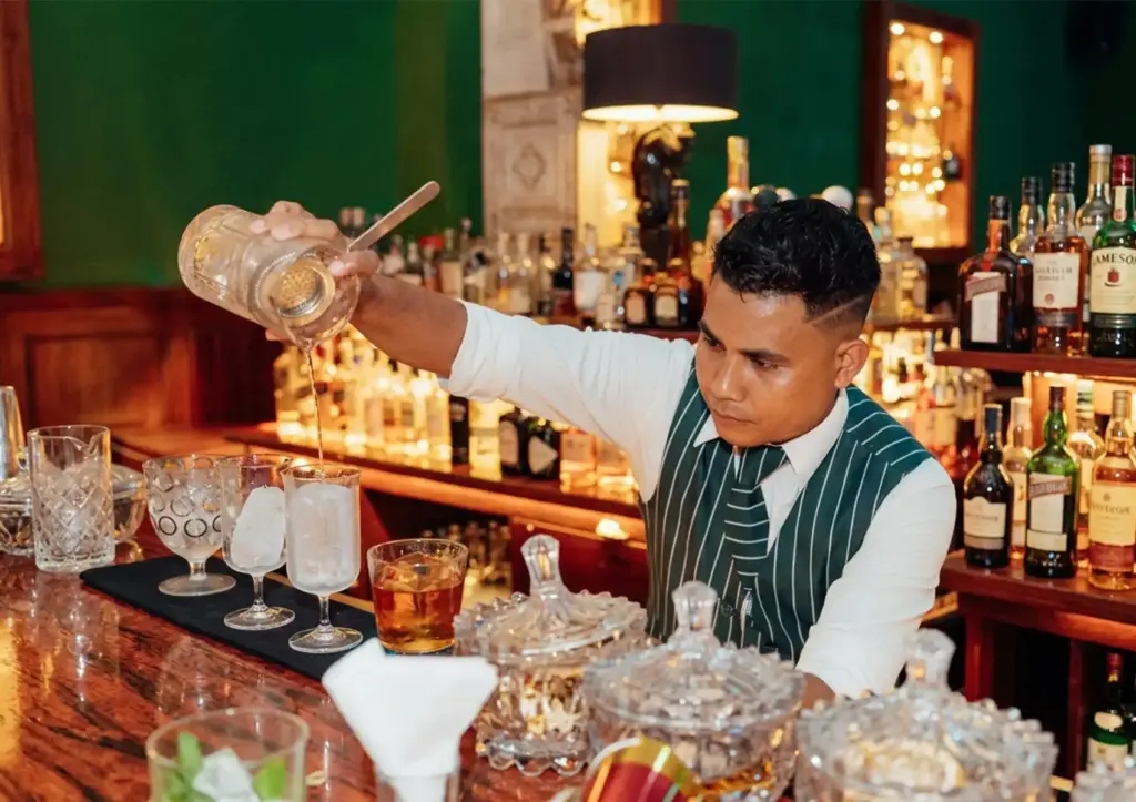 A bartender wearing a green striped vest pours a mixed drink from a mixing glass into an ice-filled glass at a cocktail bar ubud.