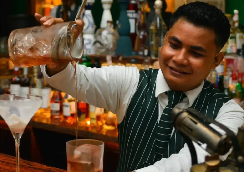 A smiling bartender in a green striped vest pouring a drink from a mixing glass at a cocktail bar Ubud.