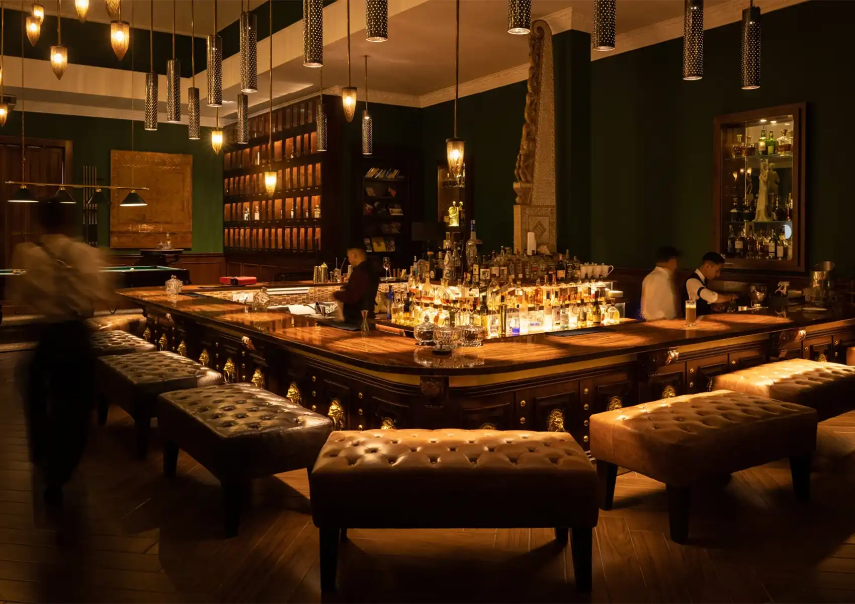 The sophisticated, dimly lit interior of a speakeasy-style cocktail bar ubud, featuring a polished wooden counter, leather tufted ottomans, and an illuminated liquor display.