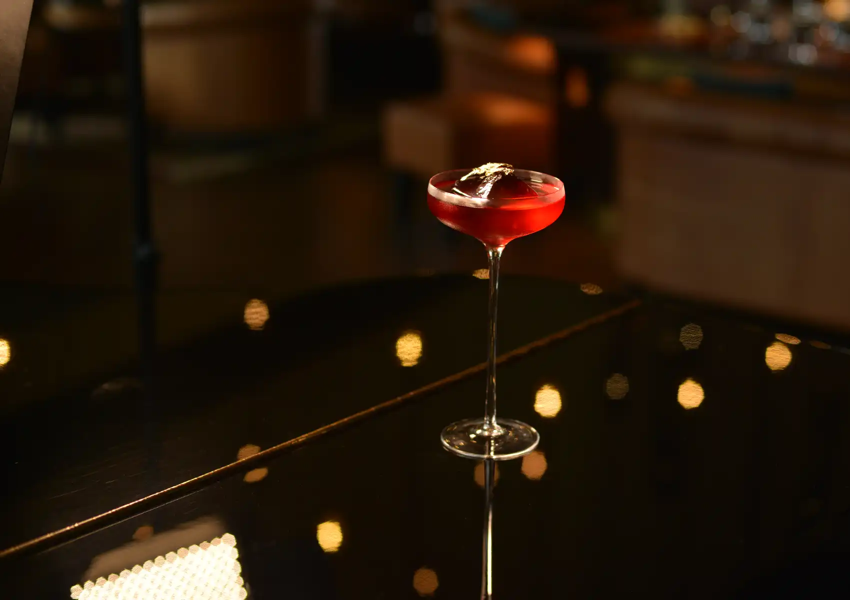 A vibrant red cocktail served in an elegant coupe glass on a dark, reflective table with warm bokeh lights at a cocktail bar ubud.