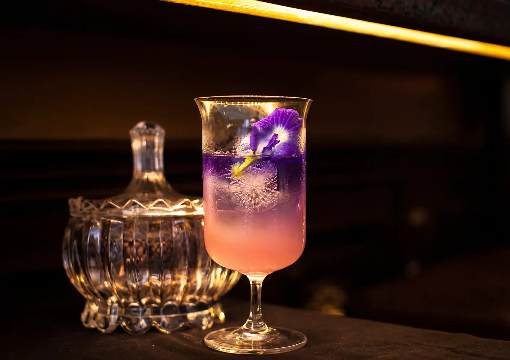 A vibrant two-toned purple and pink mezcal cocktail garnished with a flower, sitting next to a crystal jar on a counter at a cocktail bar ubud.