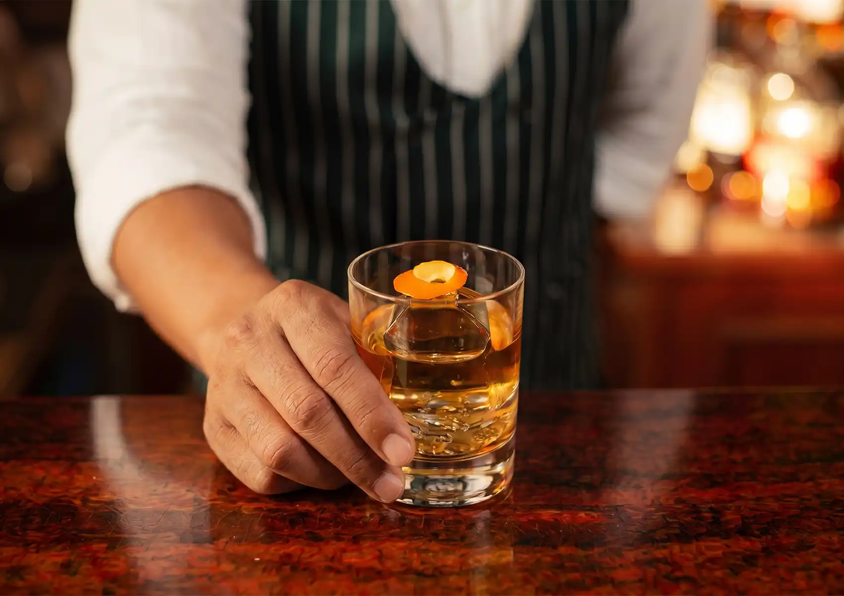 A classic Old Fashioned cocktail with a large ice cube and orange peel garnish, resting on a reflective black bar top at a dimly lit cocktail bar ubud.