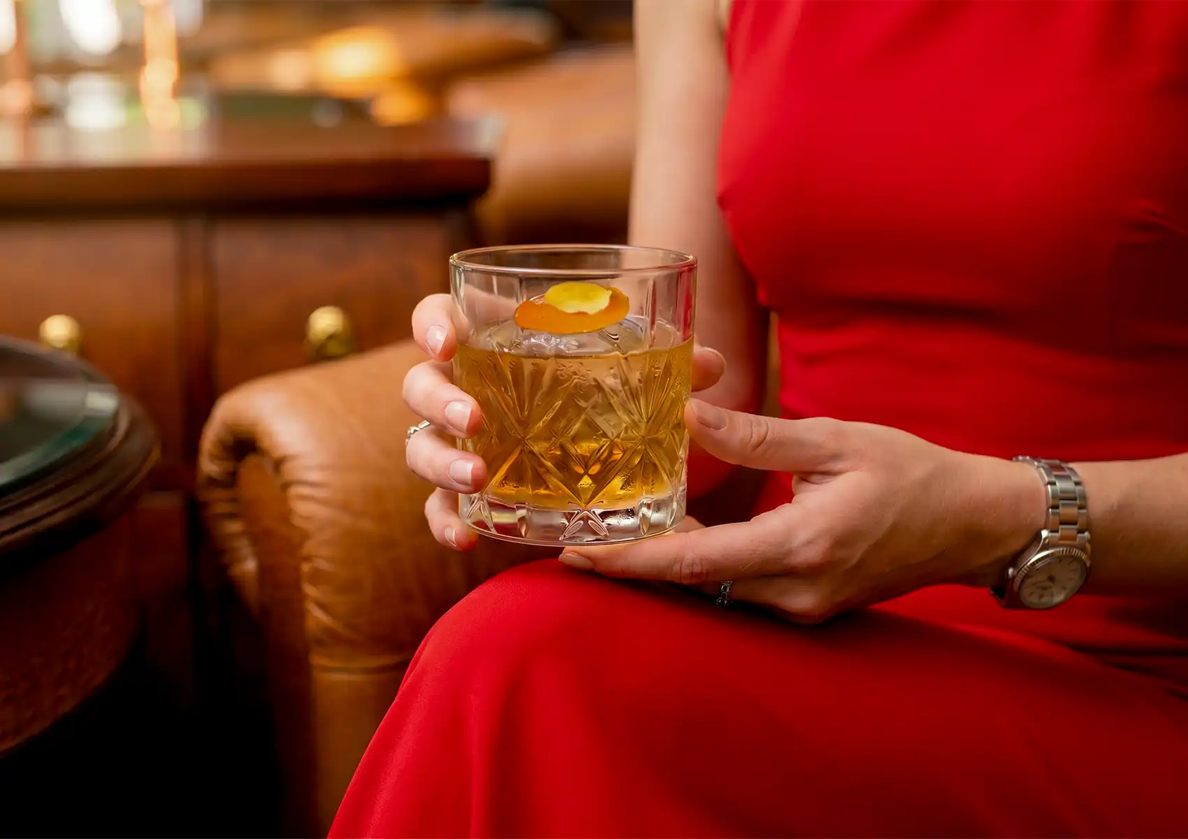 A woman wearing a vibrant red dress sits in a brown leather chair, holding a beautifully cut crystal glass containing an Old Fashioned at an elegant cocktail bar ubud.