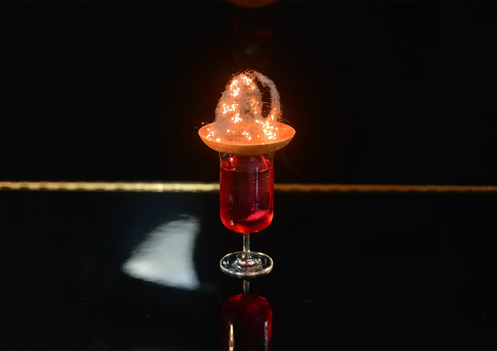 A vibrant red signature drink topped with a glowing, intricate spun garnish on a wooden rim, served in a dimly lit cocktail bar Ubud.