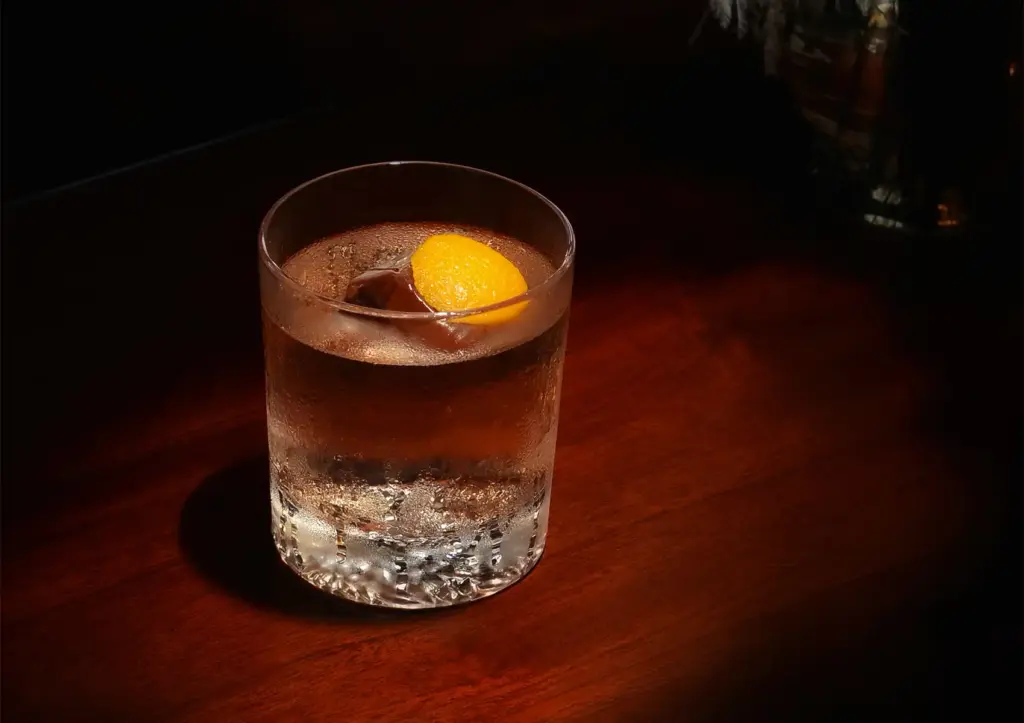 A crystal-clear signature drink served over a large, single ice cube and garnished with a bright orange twist, resting on a polished wooden counter at a premium cocktail bar Ubud.
