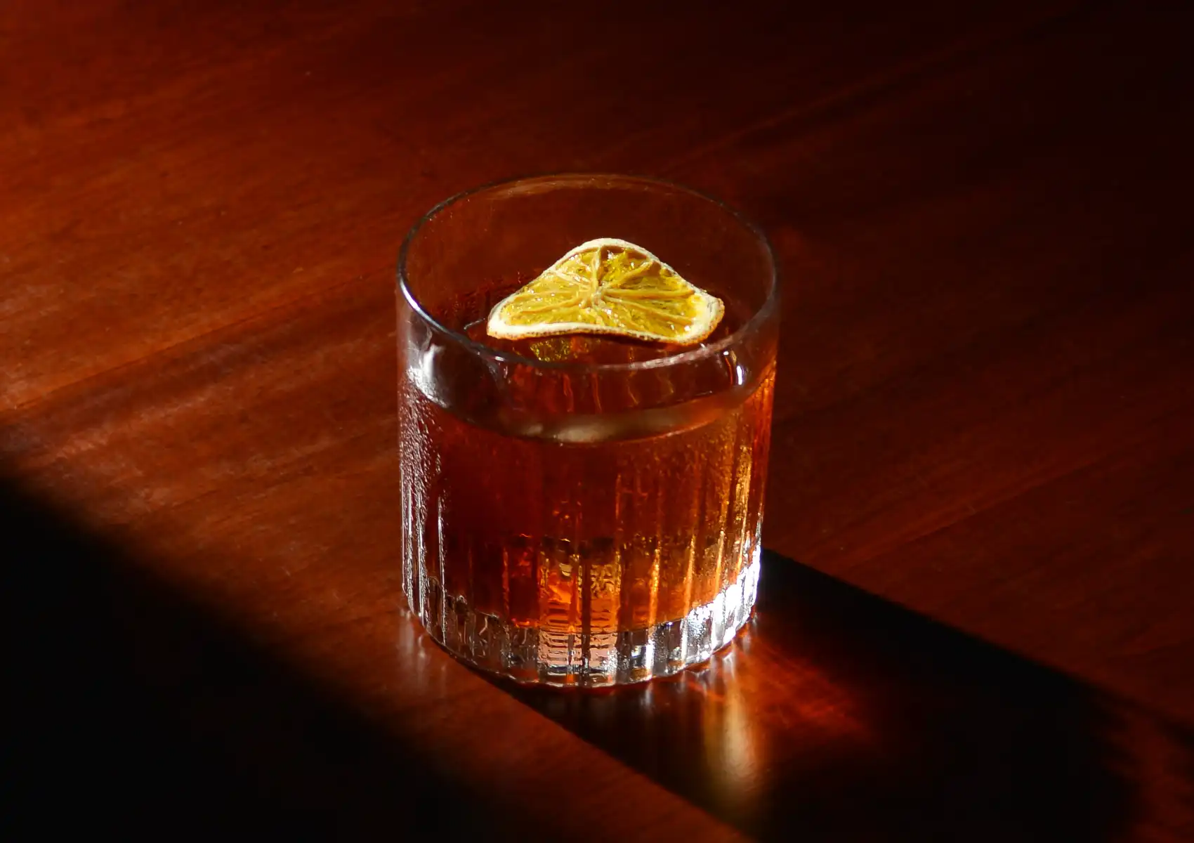 A rich, amber-colored drink garnished with a dehydrated citrus wheel in a textured rocks glass, showcased under dramatic lighting at a sophisticated cocktail bar Ubud.