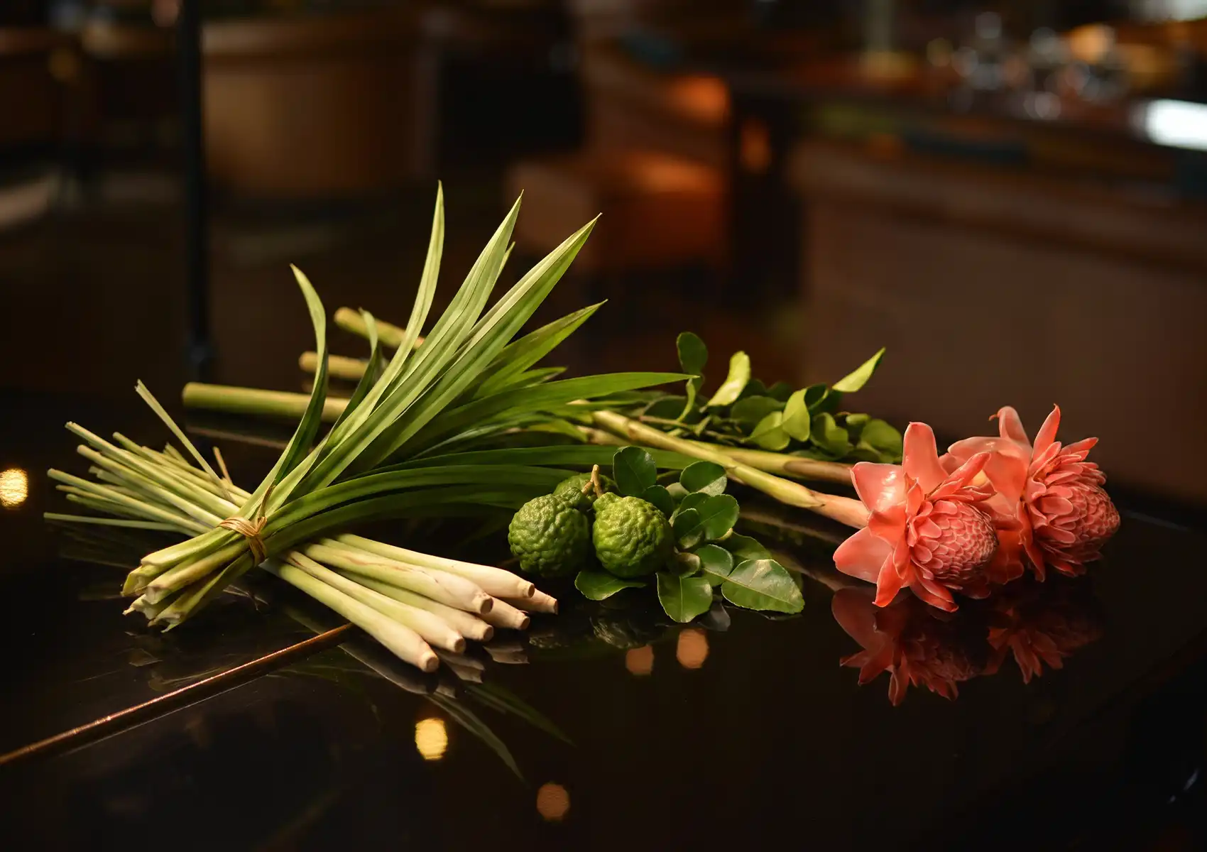 A vibrant display of fresh local botanicals, including bundled lemongrass, kaffir limes, and bright pink torch ginger flowers, resting on a dark reflective counter at a cocktail bar ubud.