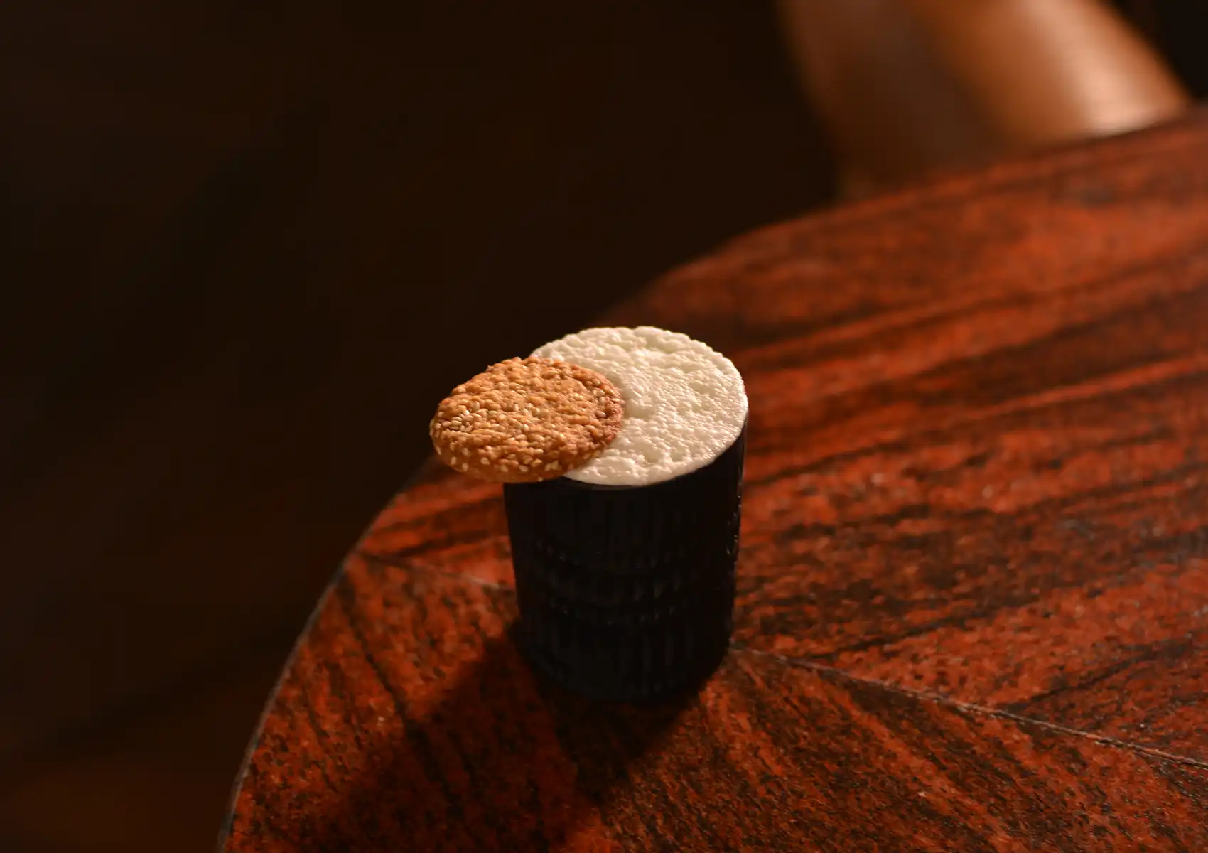 Close-up of a creative cocktail served in a textured black glass featuring a white foam top and a sesame cookie garnish on a wooden table at a cocktail bar ubud.