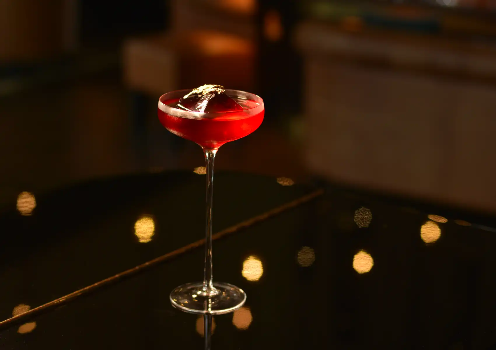 A bright red cocktail served in a classic coupe glass featuring a large ice cube with a gold leaf garnish, displayed on a sleek table at a cocktail bar ubud.