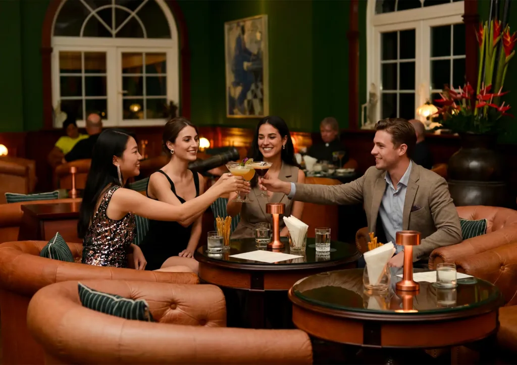 A group of smiling friends in elegant attire toasting with their drinks while relaxing in tufted leather armchairs at a cocktail bar ubud.