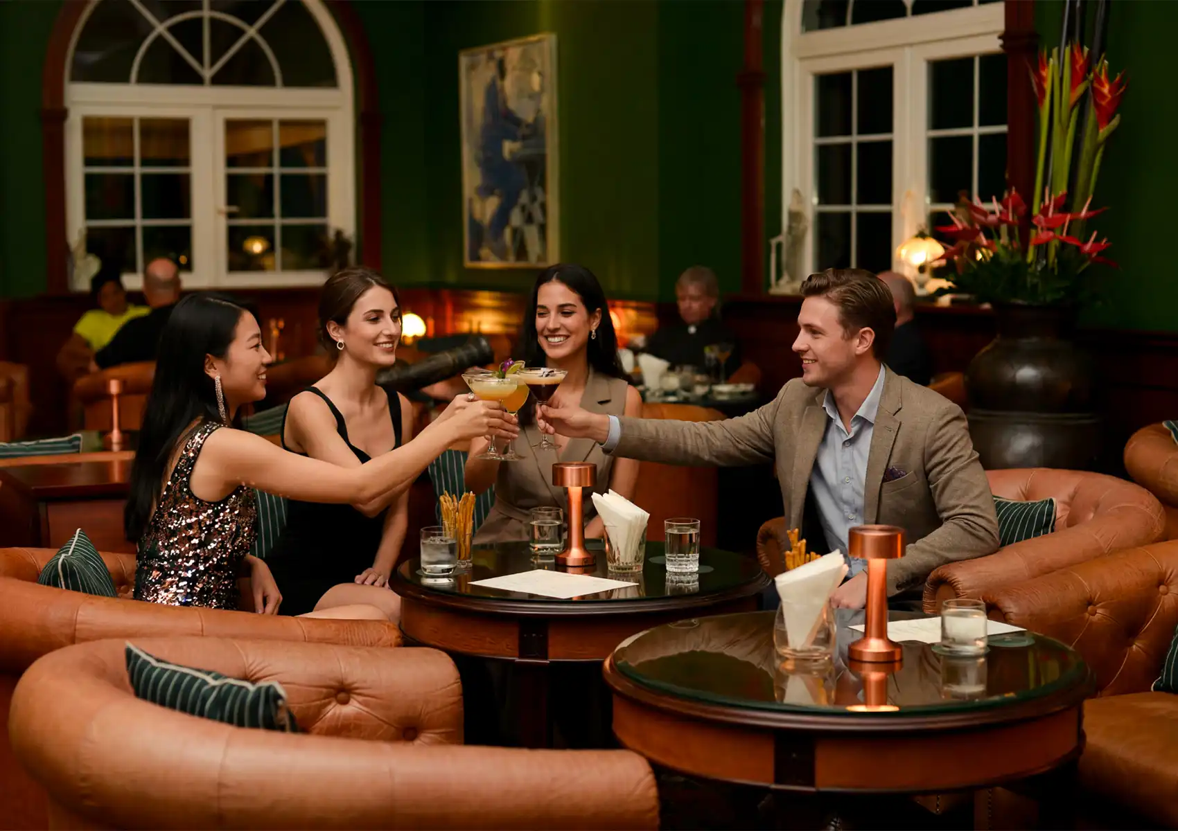 Four smiling friends toast with drinks in martini glasses while seated in leather armchairs around a glass table at a cocktail bar ubud.