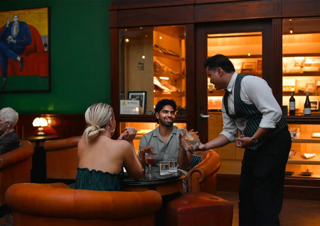 A waiter in a striped vest pours a drink from a glass decanter for a couple seated in tufted leather chairs at a cocktail bar ubud.