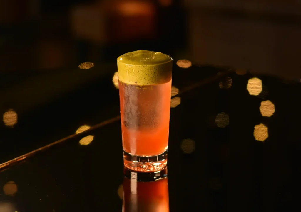 A tall pink cocktail topped with a thick layer of vibrant green foam, displayed on a dark reflective surface illuminated by warm bokeh lights at a cocktail bar ubud.