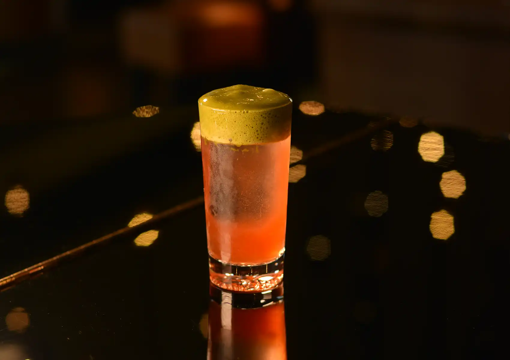 A tall pink cocktail topped with a thick layer of vibrant green foam, displayed on a dark reflective surface illuminated by warm bokeh lights at a cocktail bar ubud.