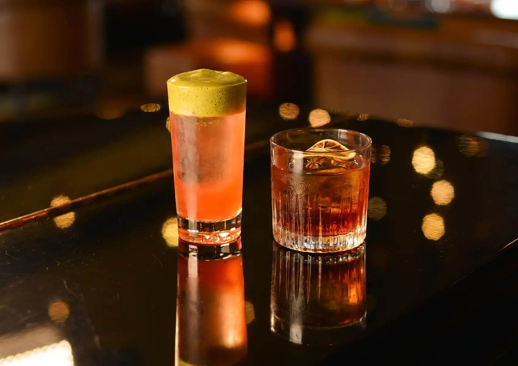 Two beautifully crafted drinks, including a tall pink beverage with green foam and a classic amber drink over a large ice cube, sitting on a dark reflective table at a cocktail bar ubud.