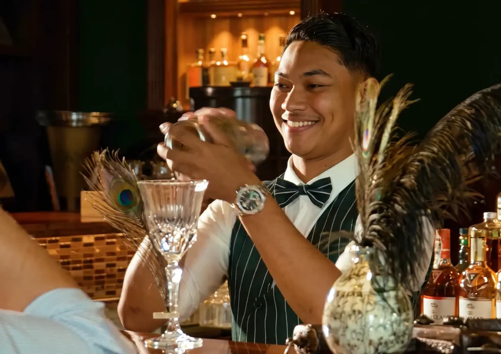 A smiling bartender in a classic bow tie and striped vest vigorously shaking a metal shaker behind the counter at a cocktail bar ubud.