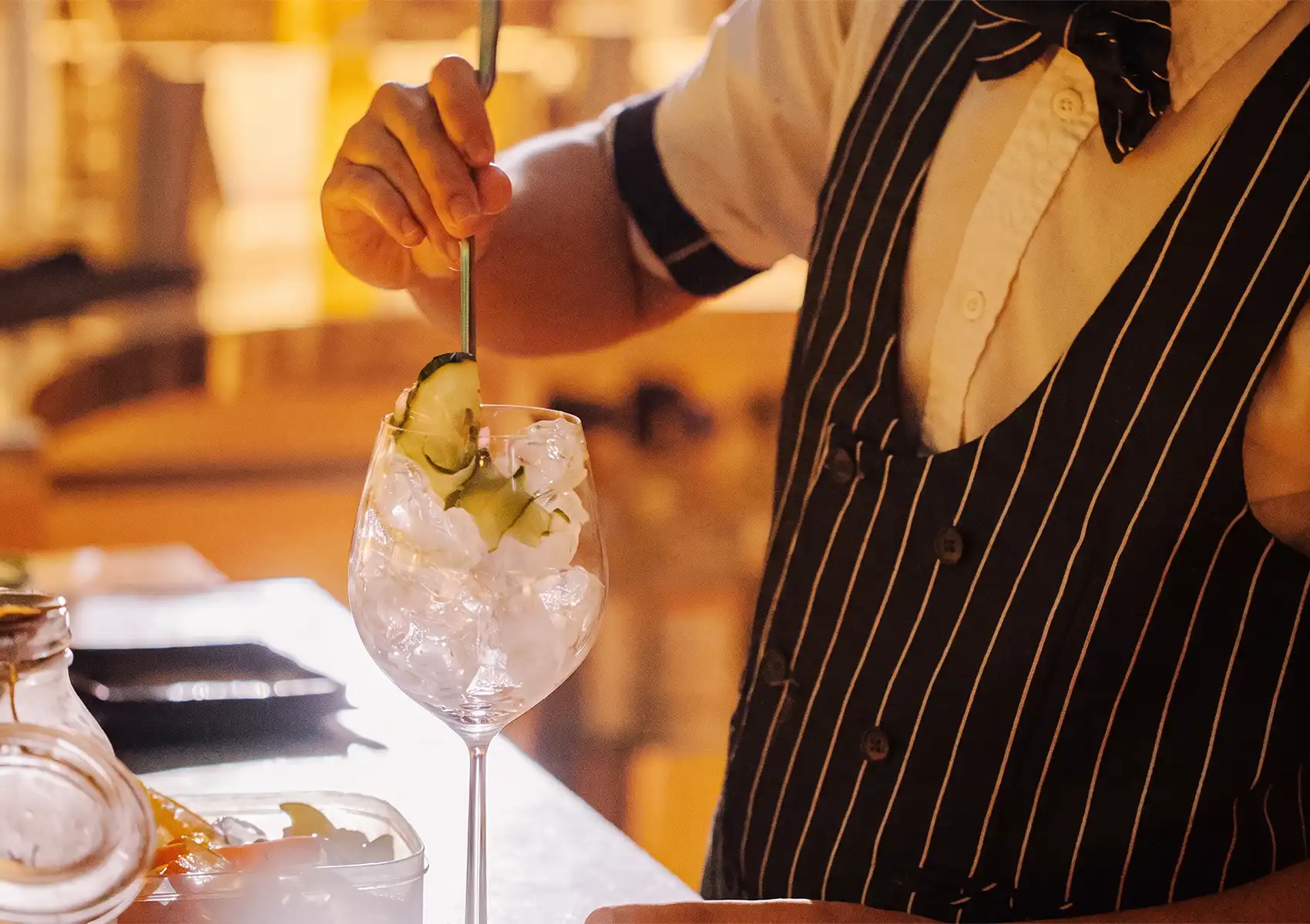 A close-up of a bartender wearing a striped vest carefully stirring a refreshing iced cocktail garnished with cucumber at a cocktail bar ubud.
