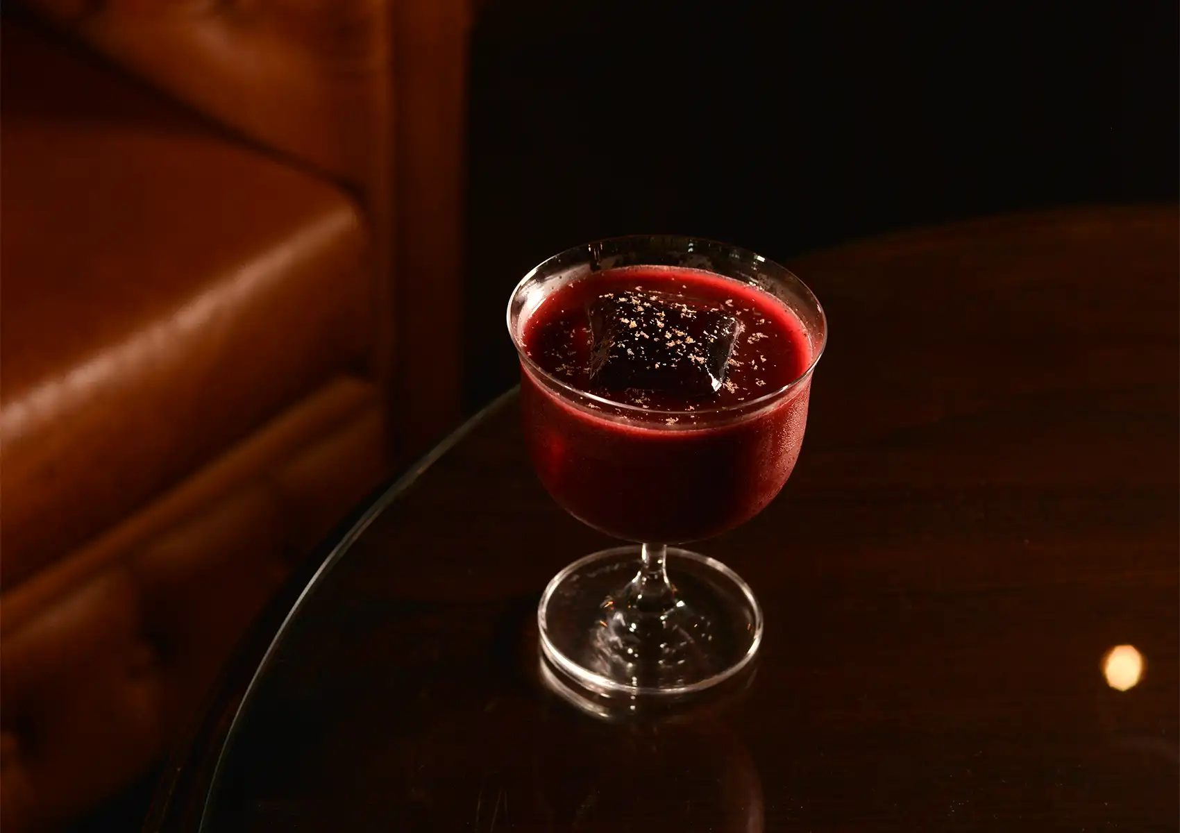 A deep red cocktail featuring a large ice cube sprinkled with delicate garnish, served in a classic glass on a dark table next to a leather armchair at a cocktail bar ubud.