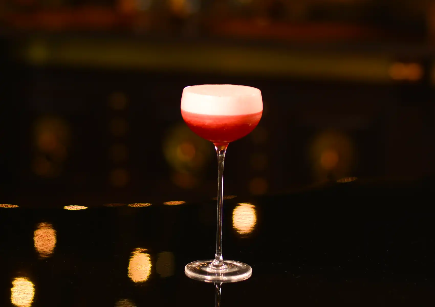 A vibrant red cocktail topped with a thick layer of white foam in a long-stemmed glass, illuminated by warm bokeh lights on a dark reflective surface at a cocktail bar ubud