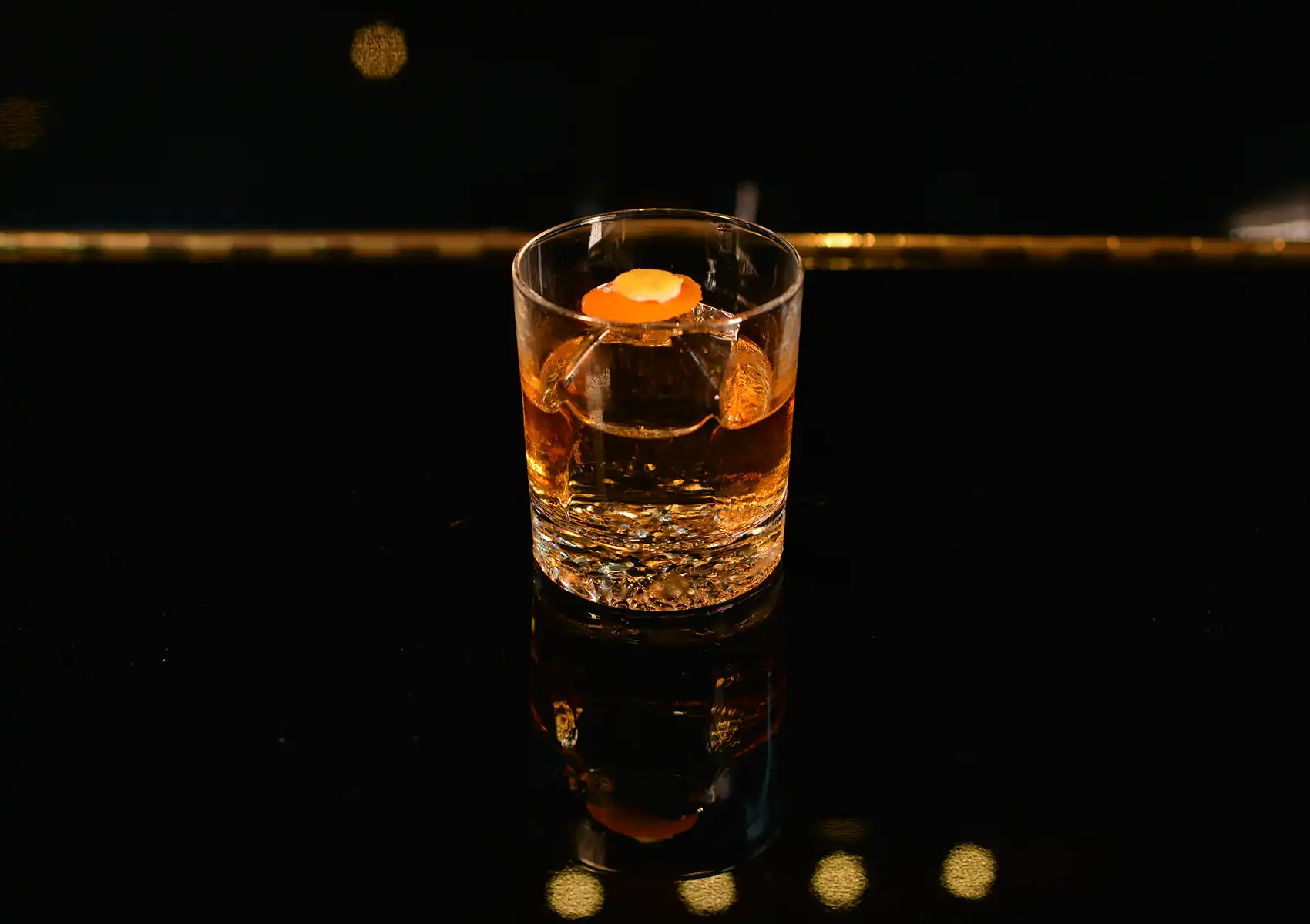A classic amber cocktail featuring a large ice cube and an orange peel garnish resting on a dark reflective counter at a stylish cocktail bar ubud.
