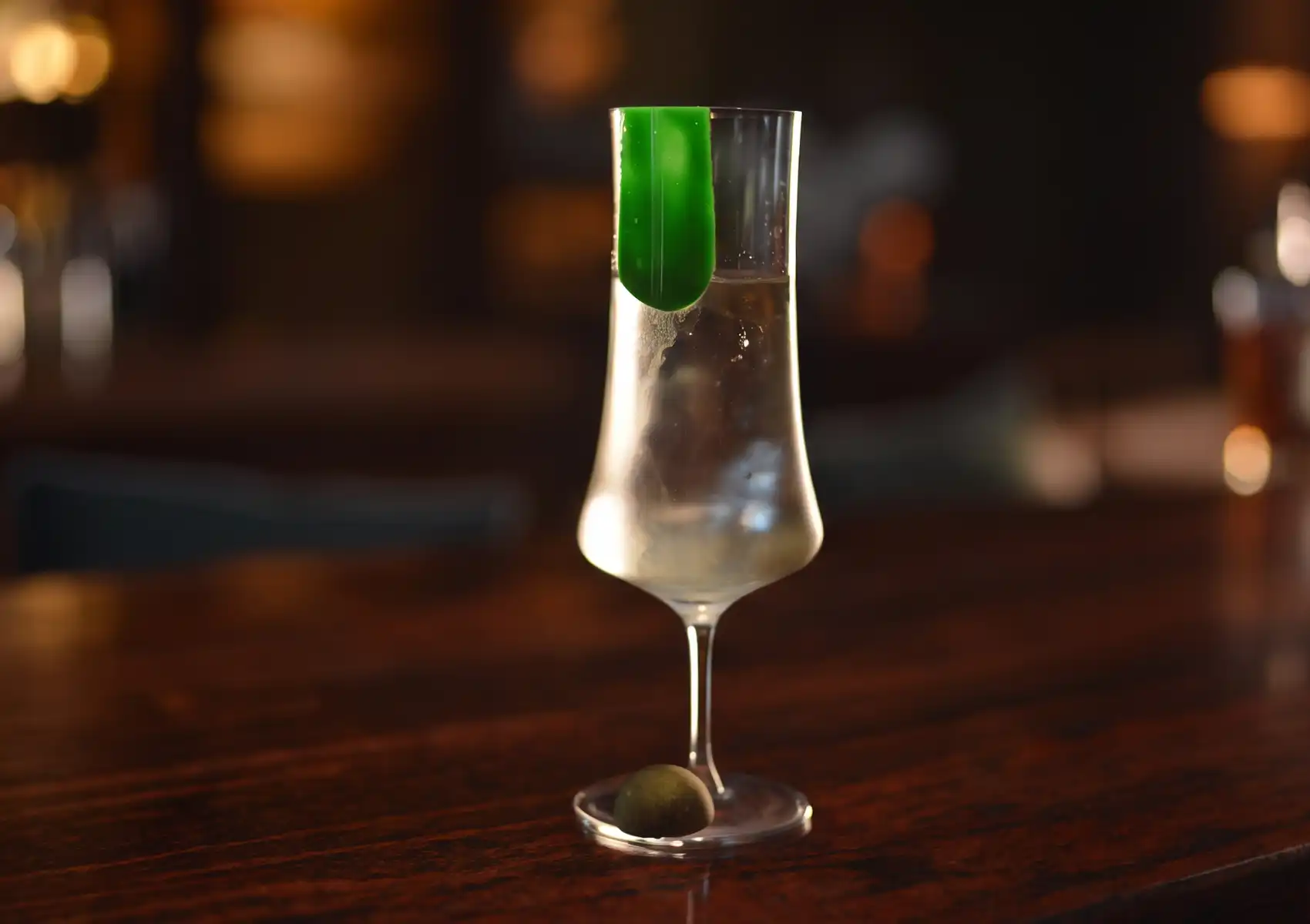 A tall, elegant clear cocktail featuring a striking green garnish inside the glass, resting on a polished wooden counter at a cocktail bar ubud.