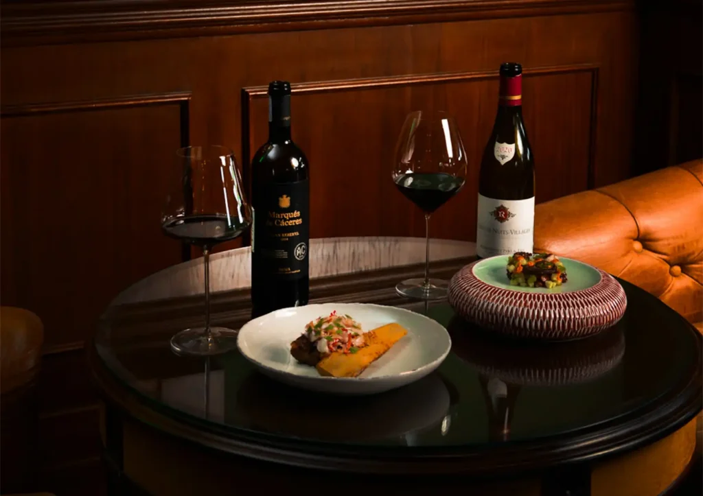 Two bottles of fine wine, filled wine glasses, and beautifully plated gourmet appetizers arranged on a round glass table next to a leather sofa at a cocktail bar ubud.