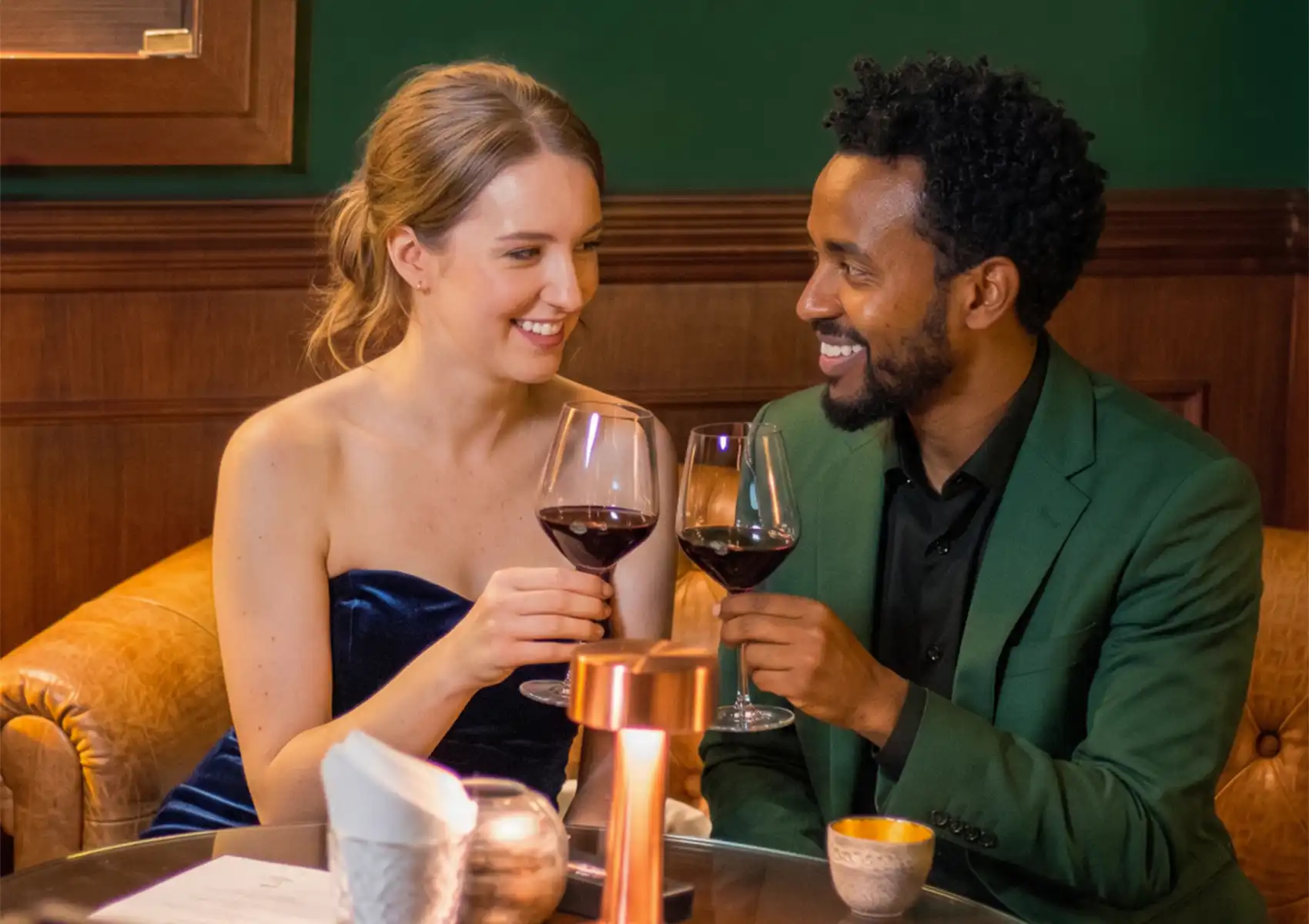 An elegantly dressed couple smiles and toasts with glasses of red wine while sitting together on a tufted leather sofa at a cocktail bar ubud.