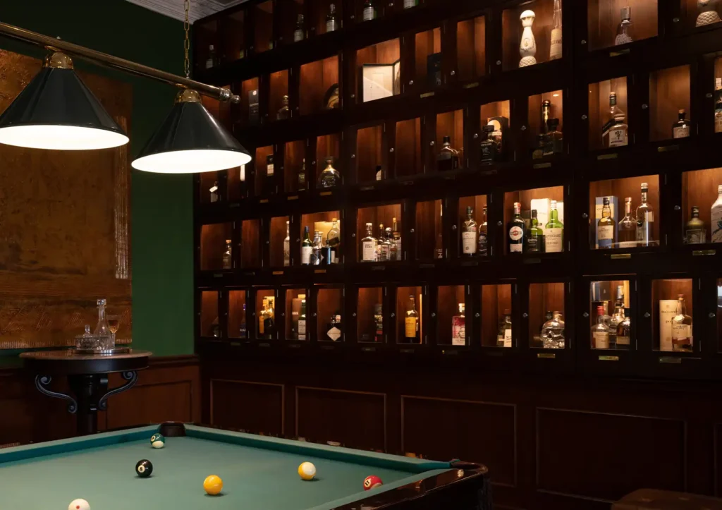 A green pool table illuminated by overhead lamps in front of a large, warmly lit wooden display cabinet filled with liquor bottles at a Cocktail bar ubud.