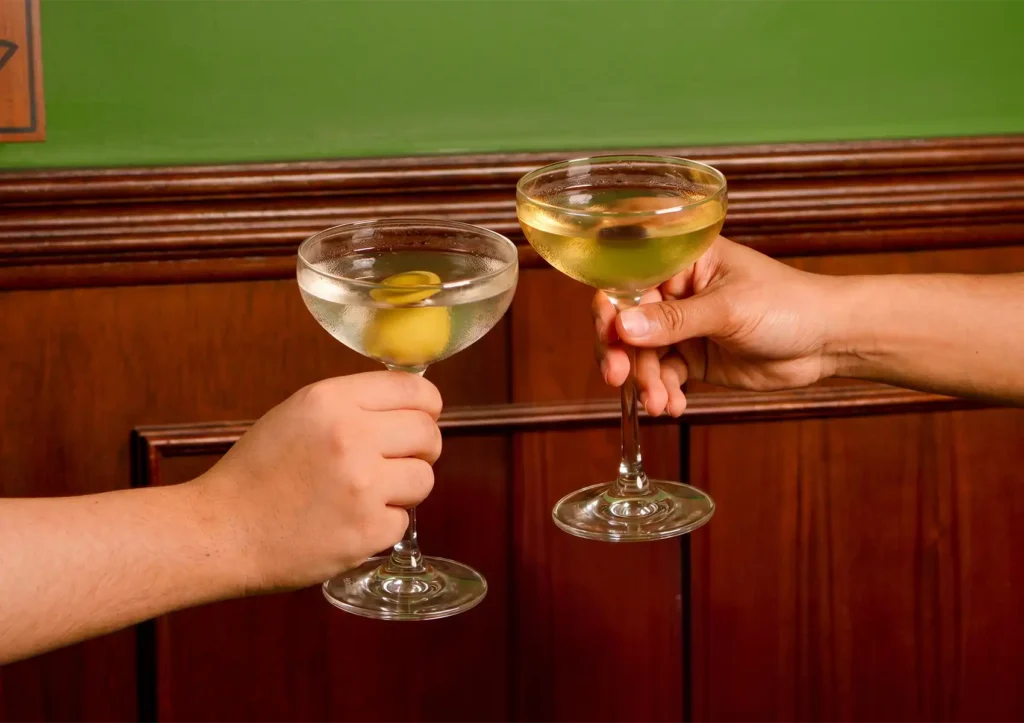 Two people clinking martini glasses garnished with olives against a wooden wall at a cocktail bar ubud.