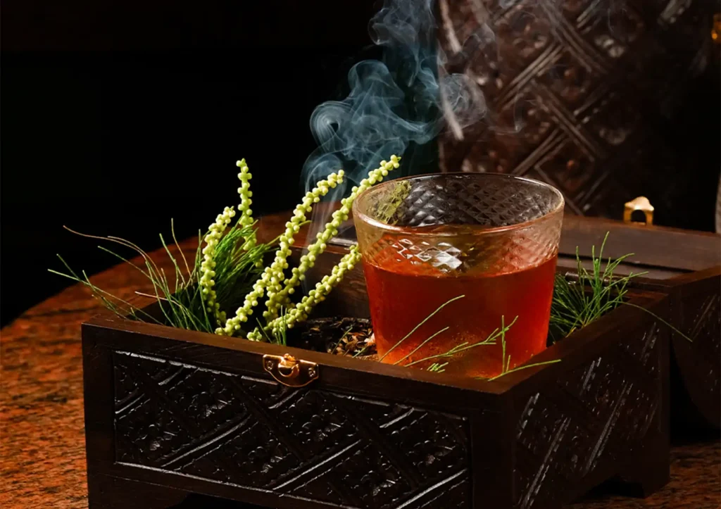 A smoky amber cocktail in a textured glass, creatively presented inside a dark, carved wooden box with fresh green botanicals at a Cocktail bar ubud.